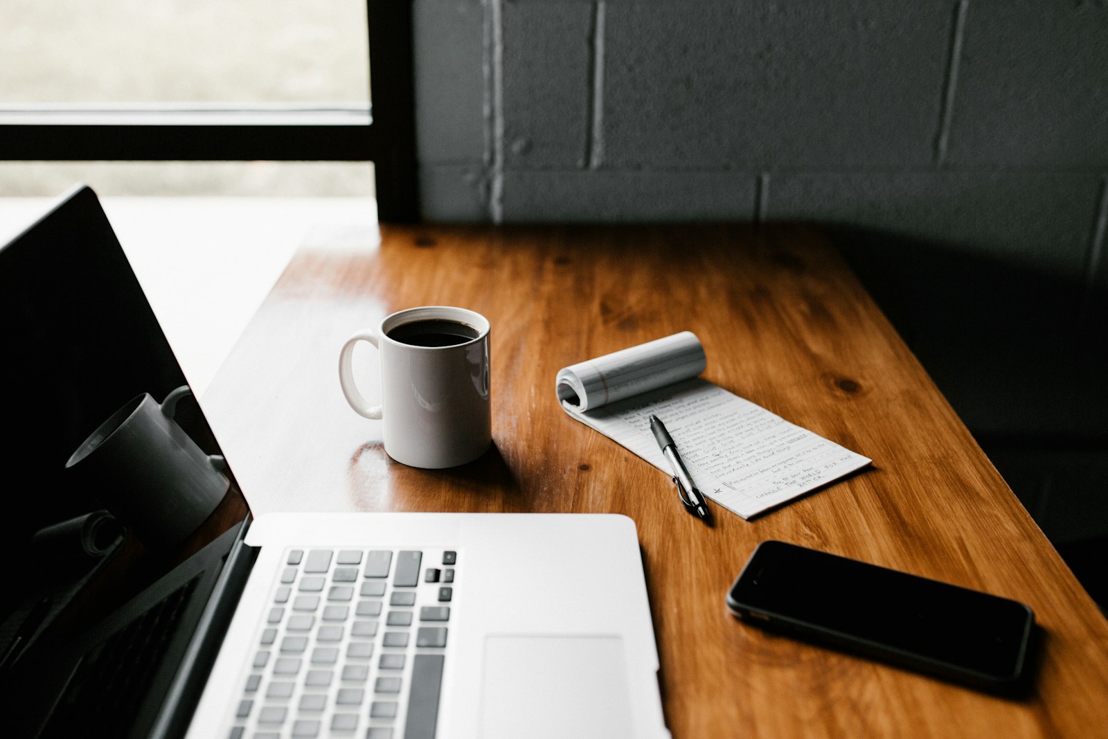 A wooden desk with a laptop, a notebook, coffee, and a phone.