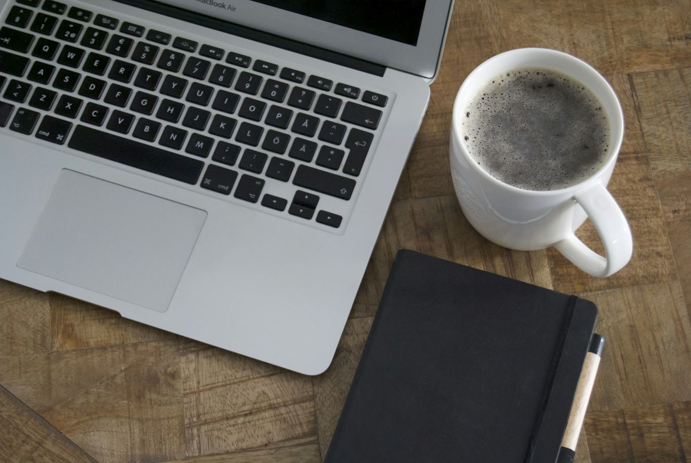 Laptop and a white mug of coffee beside a closed notebook on a wooden desk