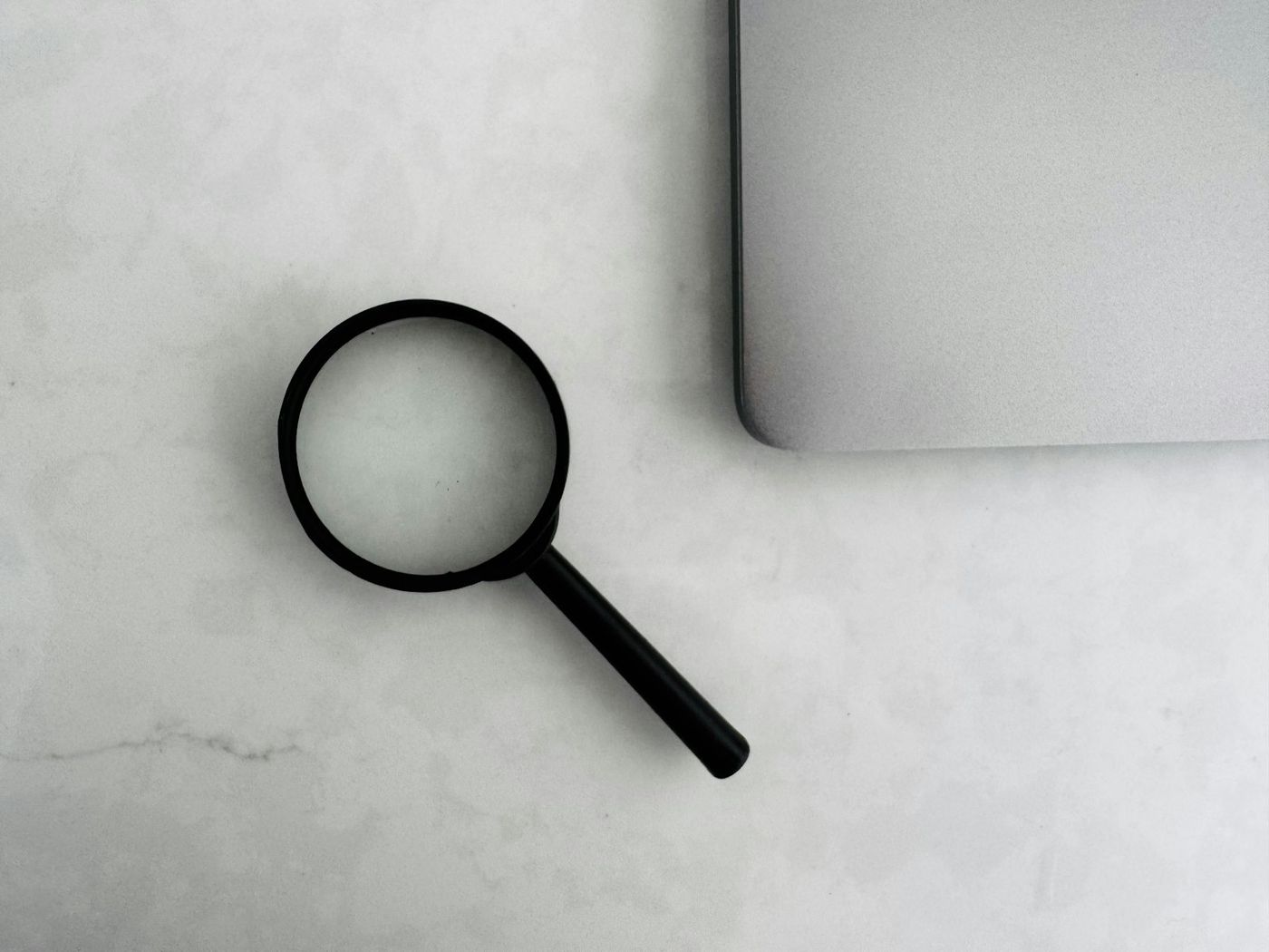 Black magnifying glass lying on a white table beside the edge of a laptop